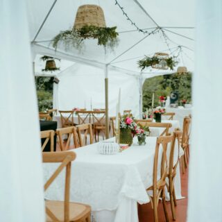 Wedding reception under a white tent with long tables, wooden chairs, and floral centerpieces on each table.