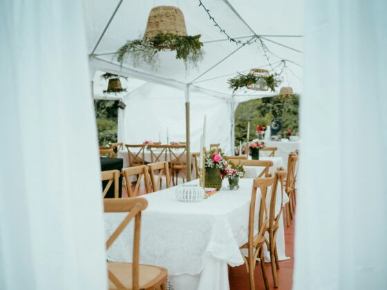 Wedding reception under a white tent with long tables, wooden chairs, and floral centerpieces on each table.