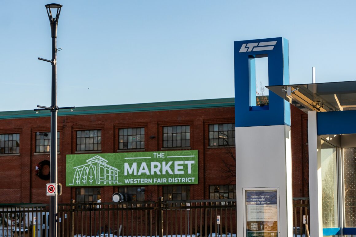 Blue architectural pillar with a white logo beside a brick building; a green sign reads 'The Market Western Fair District' near the bottom edge, with a metal fence in front.