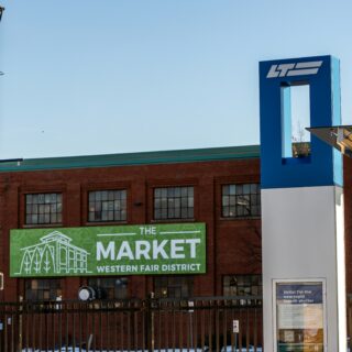 Blue architectural pillar with a white logo beside a brick building; a green sign reads 'The Market Western Fair District' near the bottom edge, with a metal fence in front.