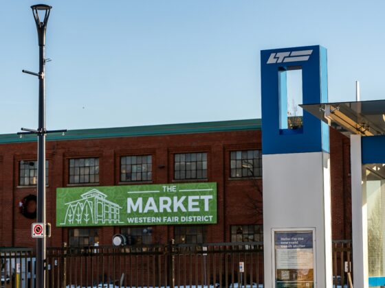 Blue architectural pillar with a white logo beside a brick building; a green sign reads 'The Market Western Fair District' near the bottom edge, with a metal fence in front.