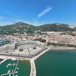 Aerial view of a harbor with numerous boats, a wide curved white building, and a stone breakwater along turquoise water with a hillside town behind.