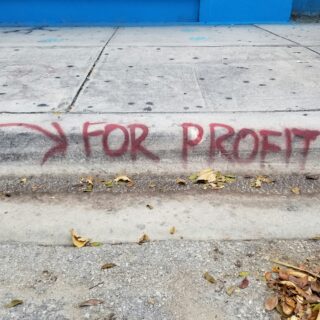 Concrete sidewalk with red graffiti letters near the curb, blue wall in the background, and fallen leaves on the ground.