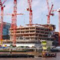 Construction site with multiple red cranes towering over a partially built building beside a river, a small boat in the foreground.