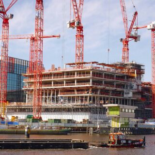 Construction site with multiple red cranes towering over a partially built building beside a river, a small boat in the foreground.