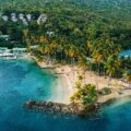 Aerial view of a tropical coastline with palm trees, sandy beach, boats, and hillside villas vs. dense greenery beneath.