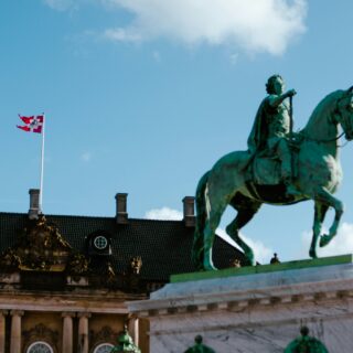 Equestrian statue of a rider on a horse atop a historic ornate building, with a Danish flag flying on a flagpole against a blue sky.