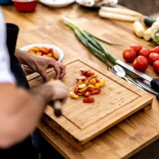 Hands chop cherry tomatoes on a wooden cutting board, with peppers and green onions nearby on a kitchen counter. Nearby are a grater and other vegetables.