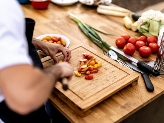 Hands chop cherry tomatoes on a wooden cutting board, with peppers and green onions nearby on a kitchen counter. Nearby are a grater and other vegetables.