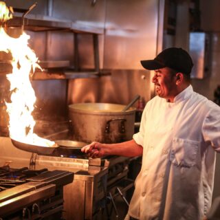 Chef in a white coat and black cap flambéing a pan on a gas stove in a busy commercial kitchen.