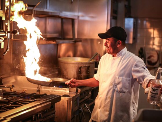 Chef in a white coat and black cap flambéing a pan on a gas stove in a busy commercial kitchen.