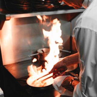 Cook in a white uniform tending to an open flame on a stove, using tongs to handle food on a plate.