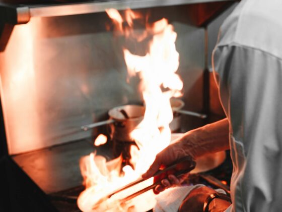 Cook in a white uniform tending to an open flame on a stove, using tongs to handle food on a plate.