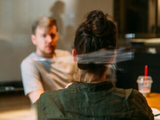 Back view of a person with dark hair tied up, wearing a green jacket, seated at a table in a cafe with another person across from them (blurry background).