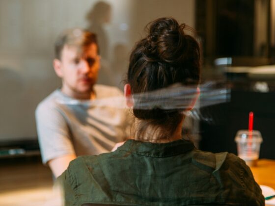 Back view of a person with dark hair tied up, wearing a green jacket, seated at a table in a cafe with another person across from them (blurry background).