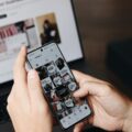 Close-up of hands holding a smartphone showing a grid of social media posts; a blurred computer monitor is in the background.