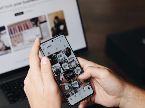 Close-up of hands holding a smartphone showing a grid of social media posts; a blurred computer monitor is in the background.