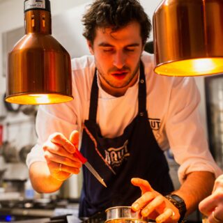 Chef in white shirt and apron plates food at a kitchen counter under warm orange pendant lights.