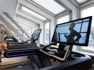 Row of modern treadmills in a bright gym, with a large display on the foreground machine showing a man running.