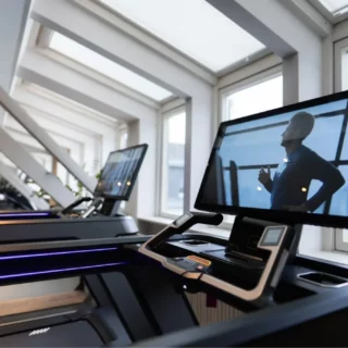 Row of modern treadmills in a bright gym, with a large display on the foreground machine showing a man running.