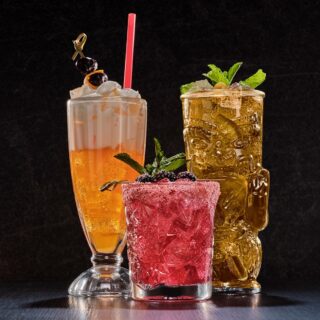 Three colorful cocktails on a dark bar backdrop: an orange drink topped with whipped cream, a pink sugared berry cocktail, and a gold tiki mug with mint.” ,