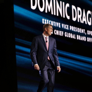 Male speaker in a navy suit and pink tie walking on a stage with a large screen displaying a name behind him.