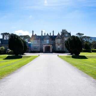Grand historic manor with stone facade at the end of a gravel driveway, flanked by manicured green lawns and tall trees under a blue sky.