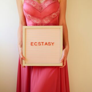 Woman in a pink lace dress holding a wooden letter board that reads ECSTASY
