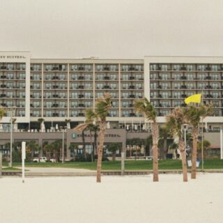 Beachfront Embassy Suites hotel seen from the sandy foreground, with palm trees and a row of balconies.