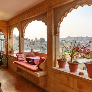 Terrace with carved wooden arches, red cushions, and potted plants overlooking a cityscape at dusk.