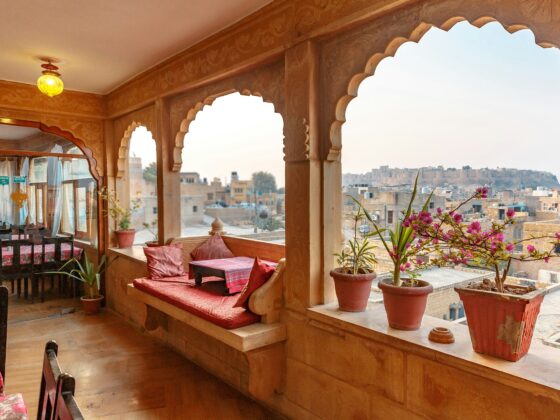 Terrace with carved wooden arches, red cushions, and potted plants overlooking a cityscape at dusk.