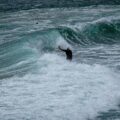 Surfer in a white helmet stands in foamy water as a large turquoise wave curls nearby.