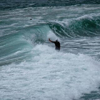 Surfer in a white helmet stands in foamy water as a large turquoise wave curls nearby.