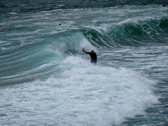 Surfer in a white helmet stands in foamy water as a large turquoise wave curls nearby.
