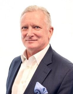 Portrait of a smiling older man in a navy check blazer and white shirt, with a light blue pocket square.