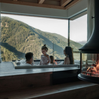 Family relaxing in a bathtub on a wooden ledge by a glass wall overlooking mountains, with a lit fireplace nearby.