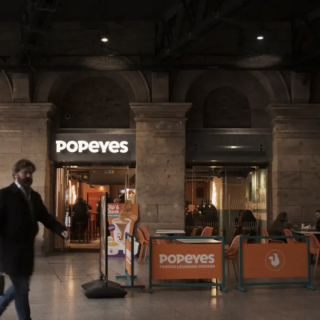 A man walks past a Popeyes restaurant inside a station, with orange barriers and outdoor seating visible outside the entrance.