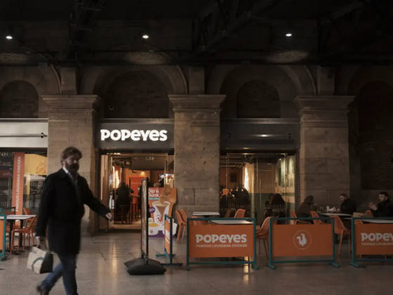 A man walks past a Popeyes restaurant inside a station, with orange barriers and outdoor seating visible outside the entrance.