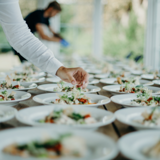 Caterer arranging salads on a long banquet table filled with white bowls in a bright, well-lit room.