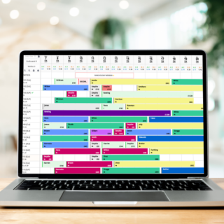 Open laptop on a wooden table displaying a colorful weekly schedule calendar on screen in a bright office setting.