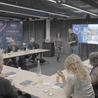 Group of professionals in a modern conference room listening to two presenters at the front with a large screen behind them and posters in the background.