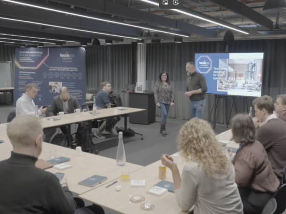 Group of professionals in a modern conference room listening to two presenters at the front with a large screen behind them and posters in the background.