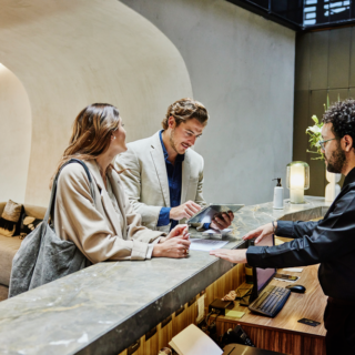 Two guests check in at a marble hotel reception as a staff member helps them with a tablet.