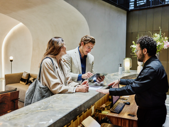 Two guests check in at a marble hotel reception as a staff member helps them with a tablet.