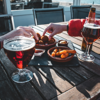 People reach for fried bites on a wooden outdoor table beside two glasses of beer by the water.