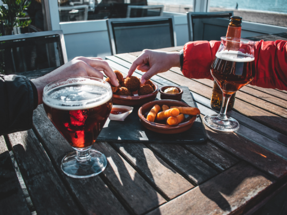 People reach for fried bites on a wooden outdoor table beside two glasses of beer by the water.