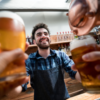 Smiling bartender in a checkered shirt and apron toasting with customers holding pint glasses.