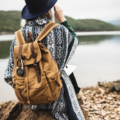 Person in a blue hat and patterned shawl sits on a rock by a lake with a tan backpack, outdoors exploration scene.