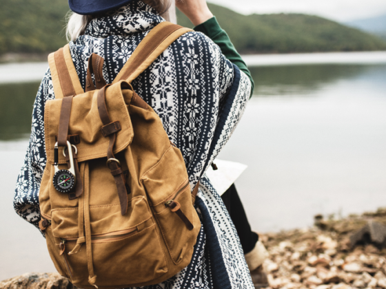 Person in a blue hat and patterned shawl sits on a rock by a lake with a tan backpack, outdoors exploration scene.
