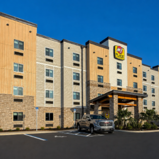 Exterior view of a My Place hotel with beige and stone façade, a covered entrance, and parked cars under a blue sky.